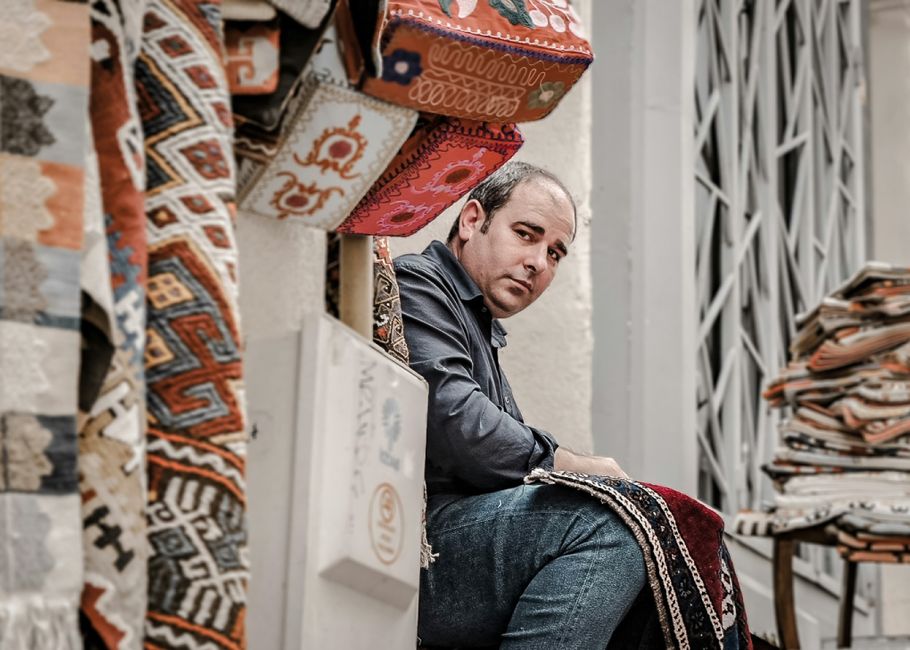 A display of intricately patterned traditional rugs in various sizes and colors, including red, orange, and beige, hung on a wooden wall in a shop or market setting. Several rolled-up rugs are stacked