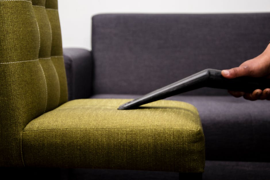 A worn, grey fabric sofa situated outdoors against a brick wall, showing visible signs of dirt and fabric wear, with a torn cushion exposing foam stuffing. The sofa's surface appears aged and stained,